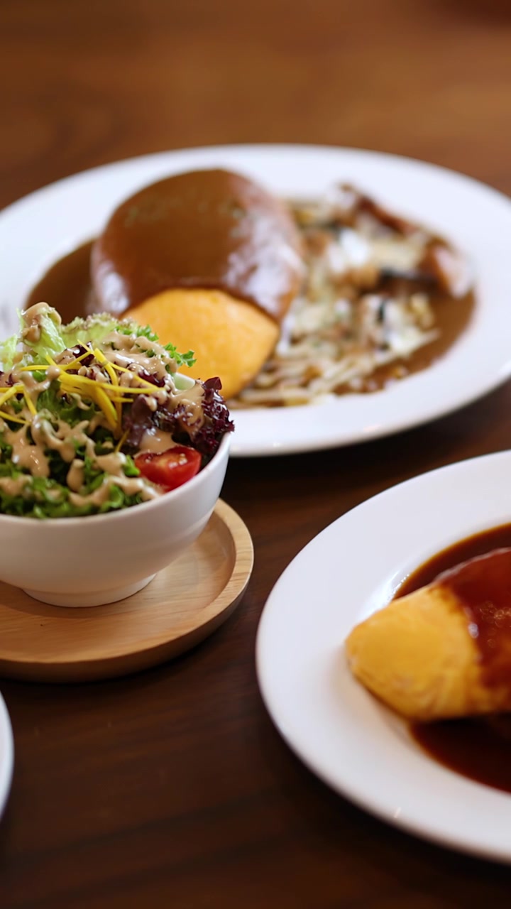 Hand places salad beside omurice and hamburg steak plate, natural light, shallow depth of field