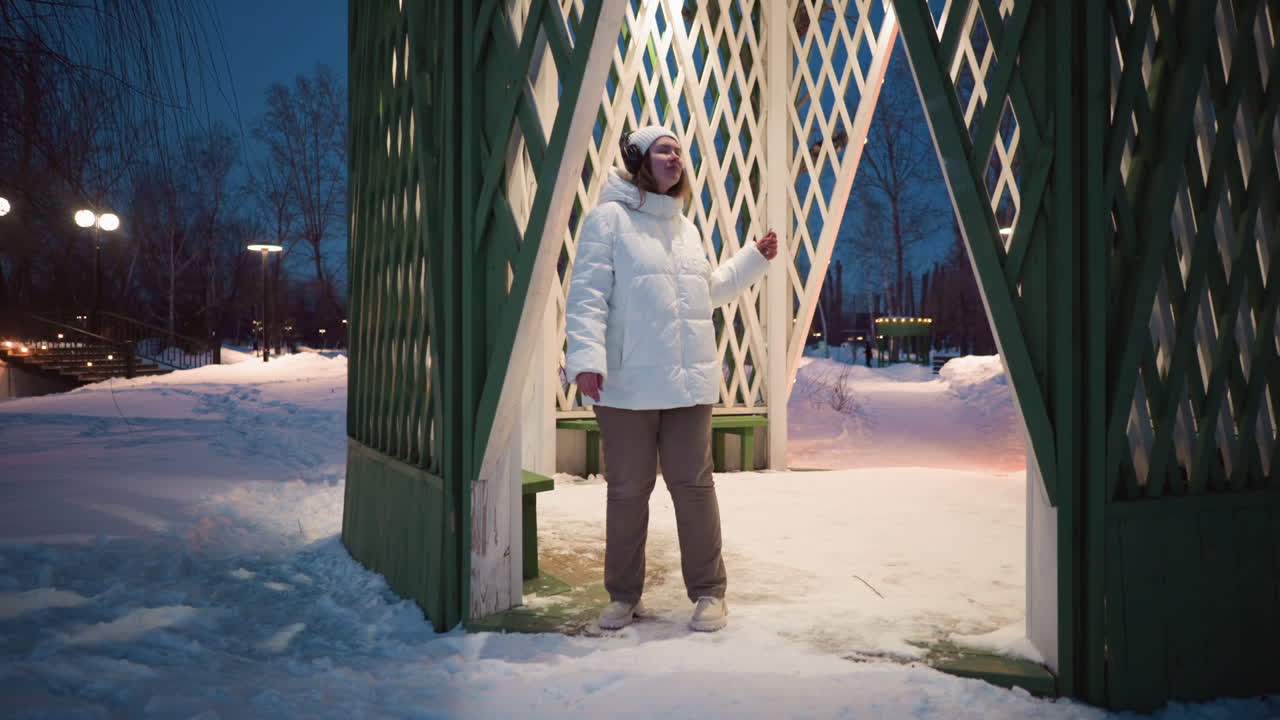 Youth wearing headphones spins at entrance of lighted gazebo in snowy park at dusk, gently moving body to rhythm under decorative winter lights and bare trees framing cozy pavilion sanctuary scene