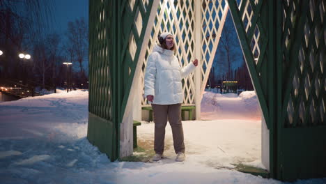 youth wearing headphones spins at entrance of lighted gazebo in snowy park at dusk, gently moving body to rhythm under decorative winter lights and bare trees framing cozy pavilion sanctuary scene
