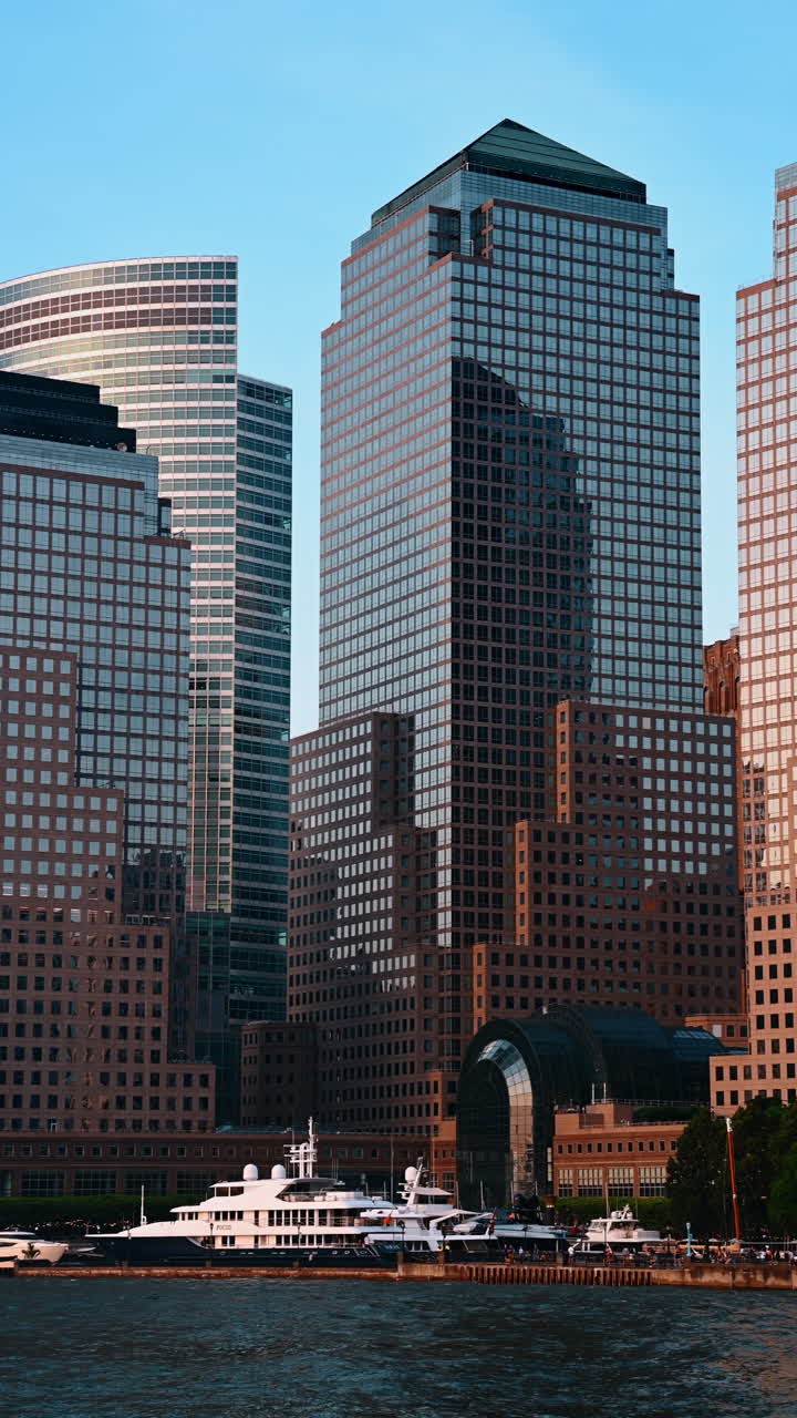 Vista vertical de imponentes arranha-céus modernos e históricos ao longo da orla de Lower Manhattan. A perspectiva próxima captura os grandes iates de luxo atracados abaixo da arquitetura comercial e residencial de grande altura