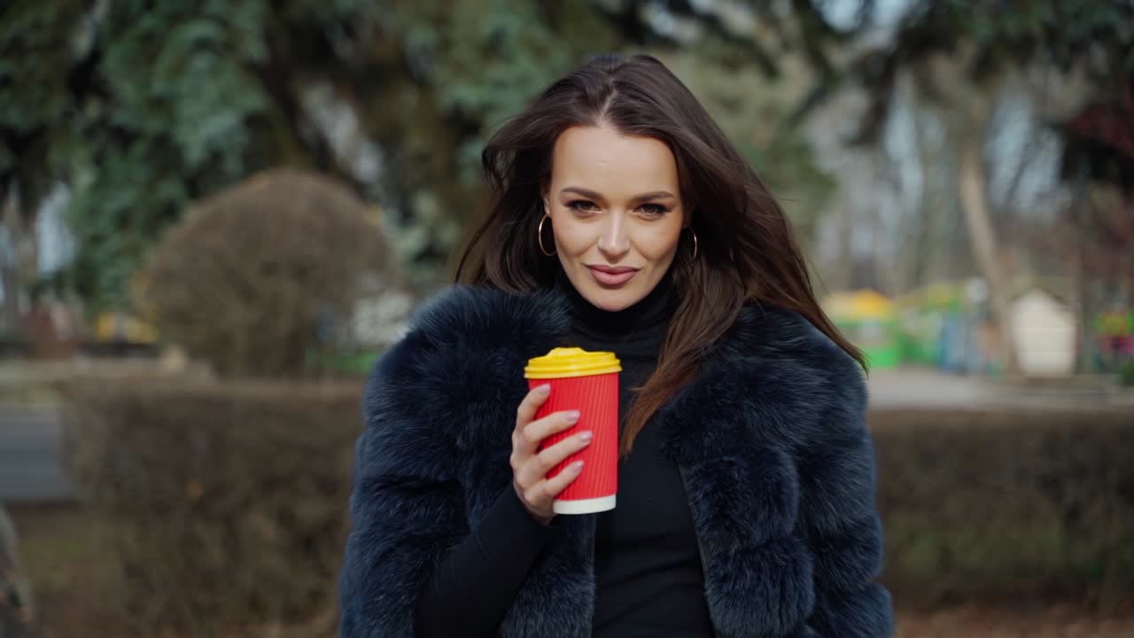 Portrait of glamorous girl with plastic coffee cup. Attractive young model in luxury fur coat standing in the park and smiling on camera. Slow motion.