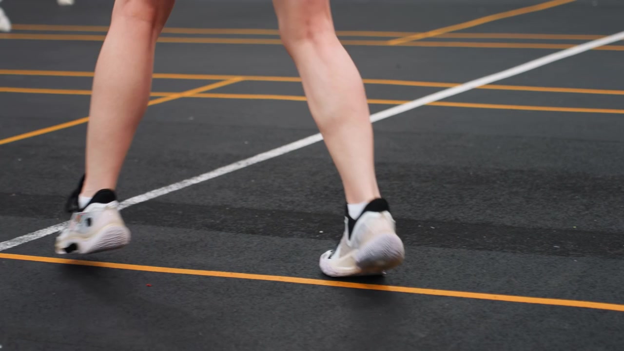 Primo piano del gioco di gambe su campo da badminton che mostra passi laterali precisi. Aderenza della scarpa da ginnastica su superficie testurizzata, linee dipinte che delimitano i confini. Atleta che pratica esercizi per equilibrio, velocità e recupero.