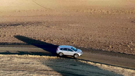 Drone-traclomg-shot-of-a-car-driving-toward-camera-along-a-narrow-dirt-road-among-golden-fields-at-sunrise