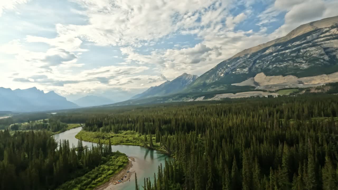 FPV drone begins at the Bow River, skimming over shimmering waters and scattered stones. It ascends gracefully, unveiling lush spruce trees, the glowing sunlit sky, and the expansive valley landscape