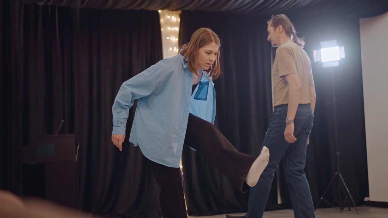 Musician coach white woman guiding student through expressive movement and vocal practice, blue shirt and casual attire, intimate coaching moment under focused lighting and dark curtains, emotional