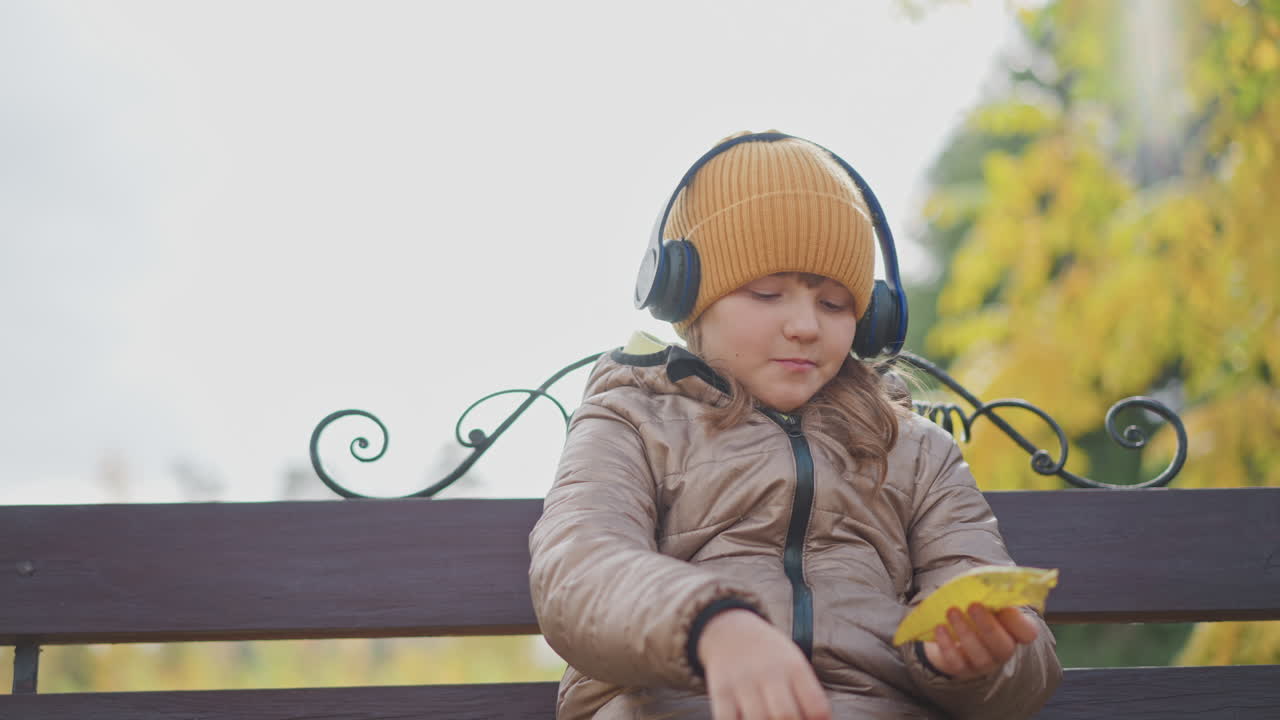 close up girl wearing beanie and headphones sitting on ornate bench in autumn park, thoughtful gaze as she examines bright yellow leaves, warm bokeh foliage backdrop and scattered leaves at her feet