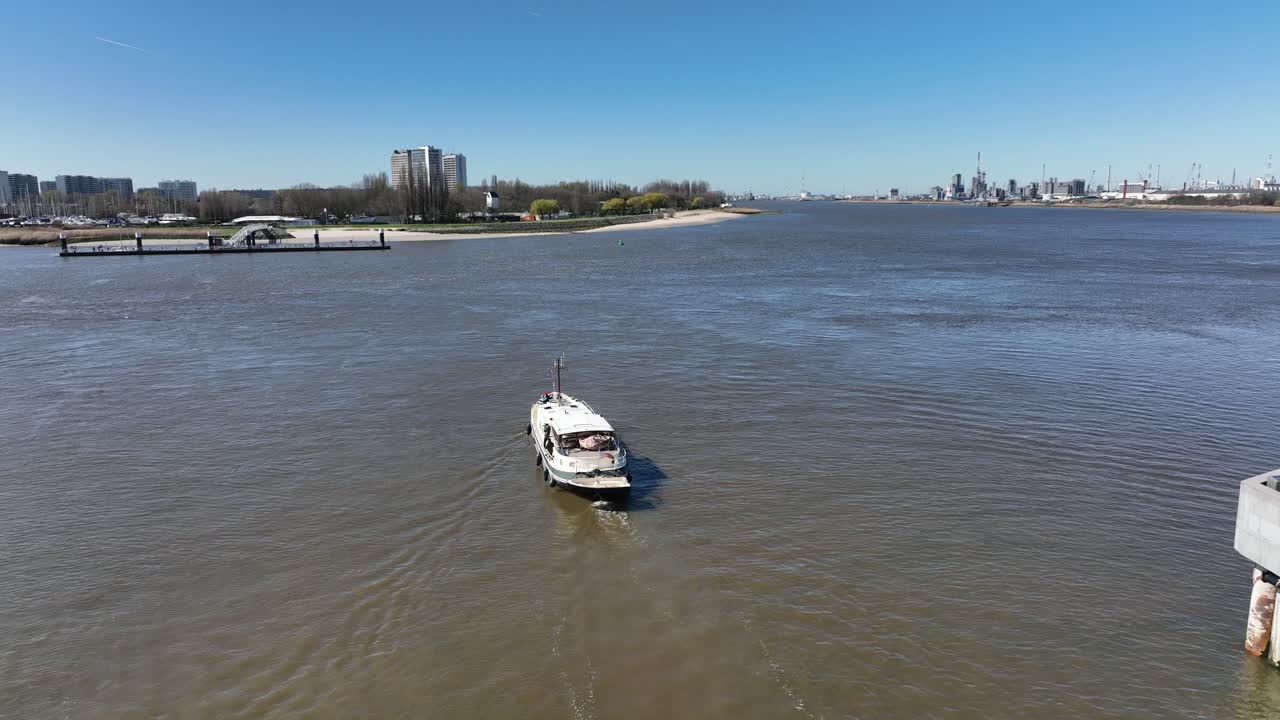 Zoom in con ripresa aerea di una barca che naviga nel fiume Anversa con torri moderne e gru portuali sullo sfondo, in una giornata limpida