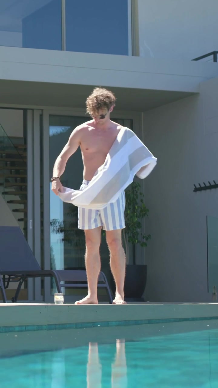 Vertical video: Exiting sliding glass door, man in striped trunks laying towel at poolside to rest