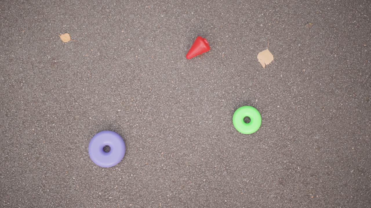 Vista dall'alto di anelli colorati e un cono sull'asfalto, foglie sparse e piccoli detriti, toni pastello tenui, tranquilla scena di vita giocosa urbana, la disposizione dei giocattoli suggerisce un gioco all'aperto casuale e delicato