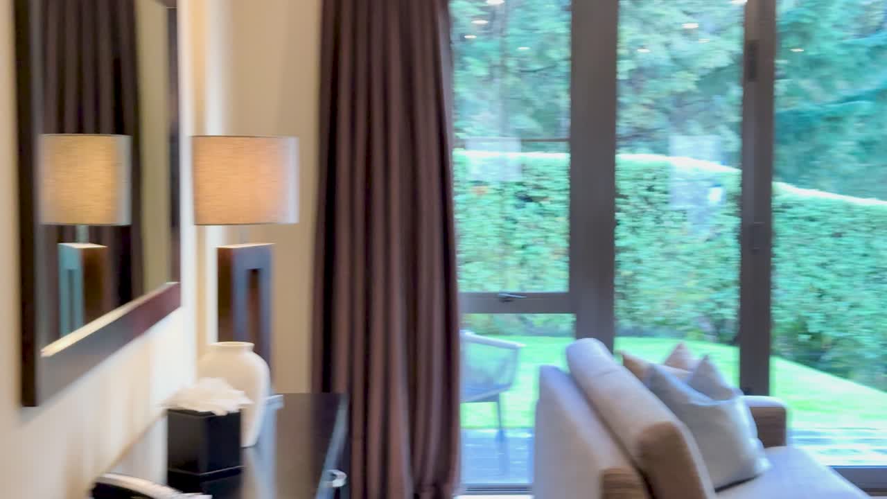 Smooth camera pan reveals a modern living room with large windows, stone fireplace, and television, bathed in warm natural daylight and soft interior lighting