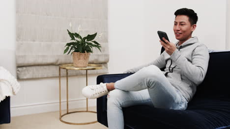 asian man relaxing on sofa, smiling while using smartphone at home