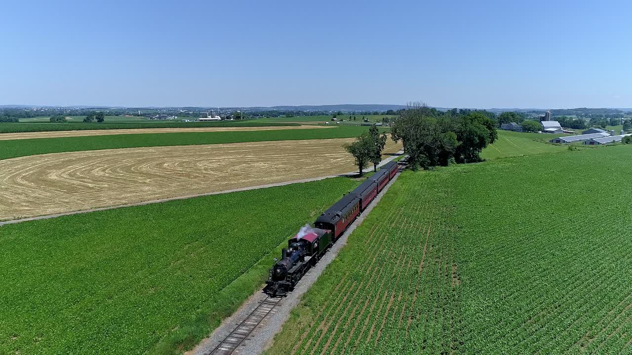 A steam train rolls through green fields and golden crops, showcasing the beauty of rural landscapes on a sunny day. Lush surroundings highlight the charm of vintage rail travel.