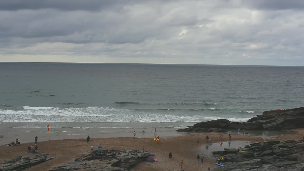 Summer day on a beach. Holiday makers enjoy their time, they swim in waters and walk around a beach. Rocky mountain is in sight. Sky is covered in clouds and ocean waters are deep blue. Footage in HD