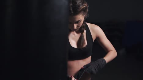 Woman-taking-on-boxing-gloves-on-hands-with-black-boxing-wraps-in-dark-room.-Slow-Motion-shot