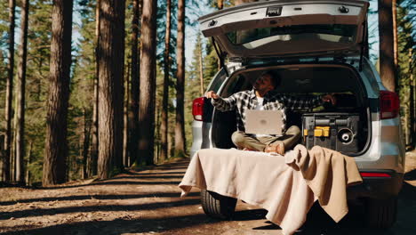 man working remotely and relaxing from car trunk in a forest