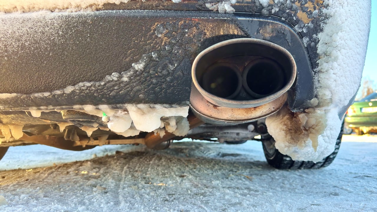 Closeup of a car exhaust pipe covered in snow and ice on a cold winter day, showing frozen road grime and frosty buildup on the rear bumper.