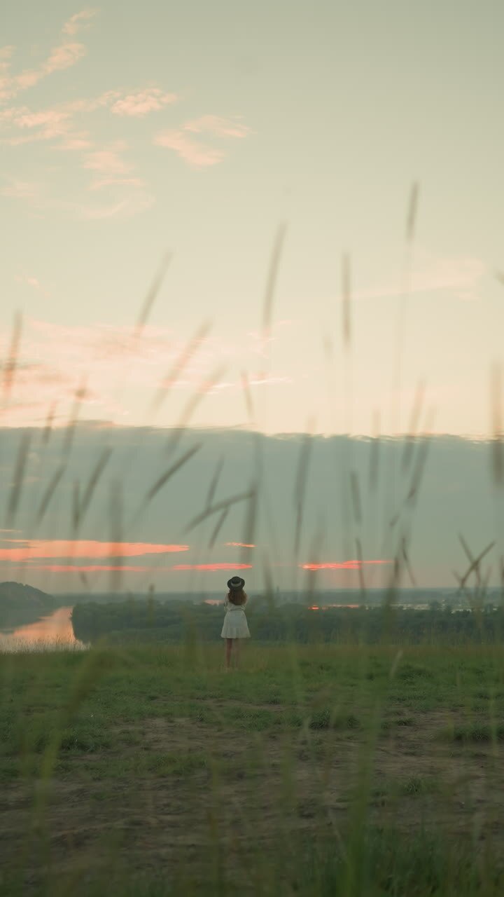 A woman in a white dress and hat stands alone in an isolated grassy field by a tranquil lake at sunset. She gazes at the serene surroundings, embracing the peaceful and reflective moment