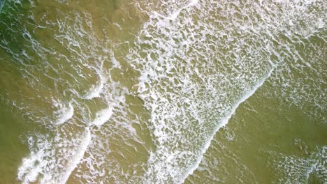 footage over shallow waves on a sandy beach. te aerial footage then raises to reveal a bigger area of waves on the weymouth beach in dorset. white crisp waves rippling over the golden sand.