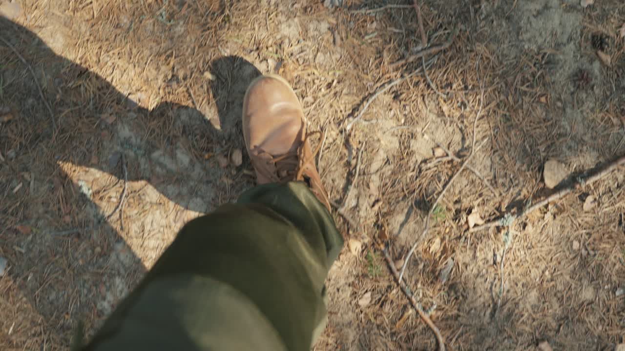 A top view of male legs in brown boots walks along a forest path covered with fallen coniferous needles.