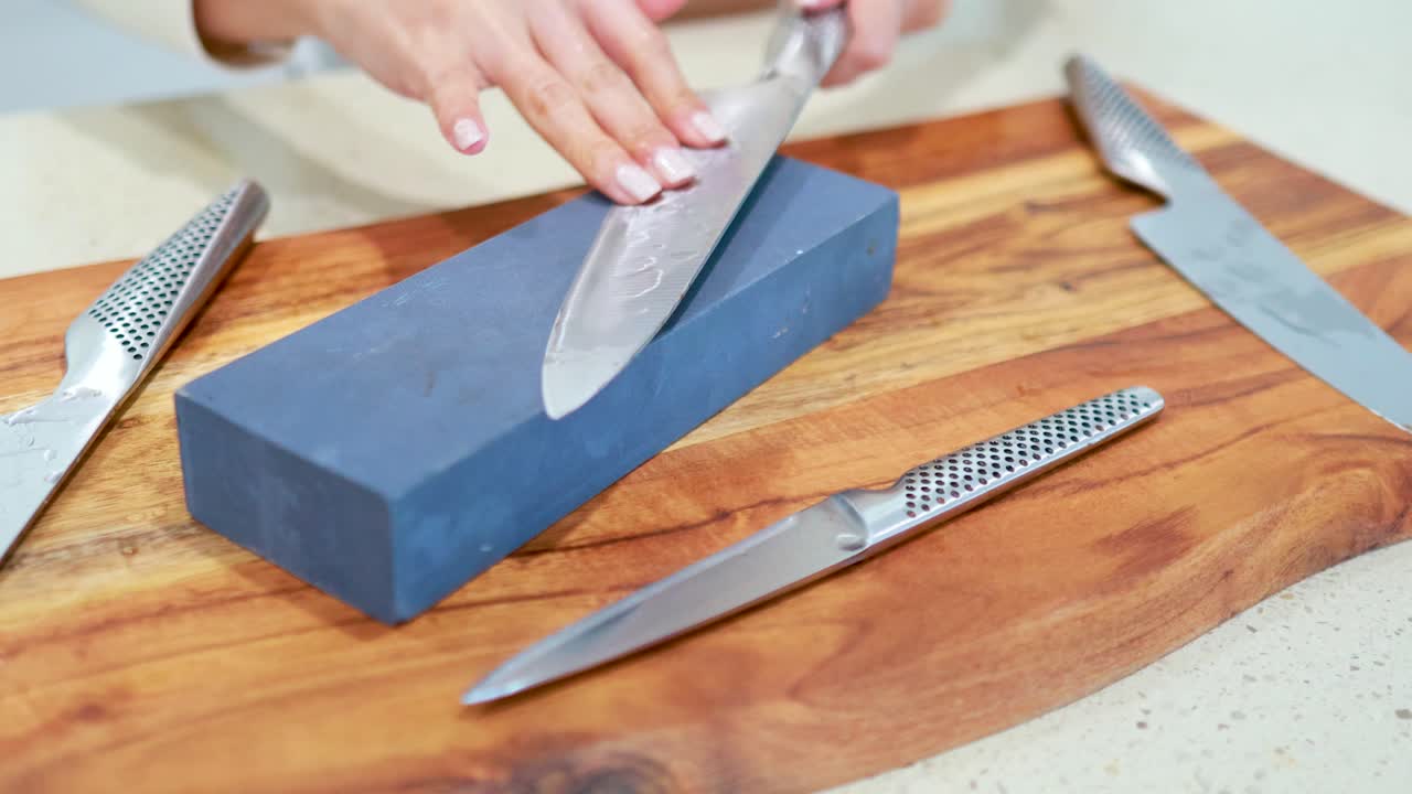 Hands sharpen a chef’s knife using a blue whetstone on a wooden cutting board under bright, even lighting with steady camera framing