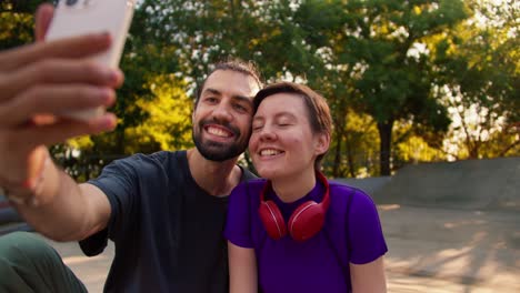 un tipo con una camiseta gris y una chica con un corte de pelo corto en una camiseta púrpura con auriculares rojos toman una selfie usando un teléfono blanco en un parque en verano