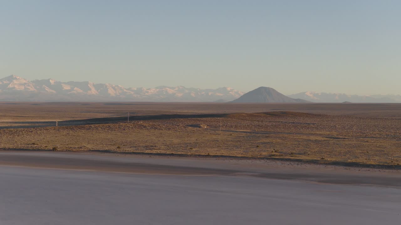 Slow drone shot going across cracked salt flats and arid fields, framing Diamante volcano and Andes in morning haze. Ideal for adventure or geology content.