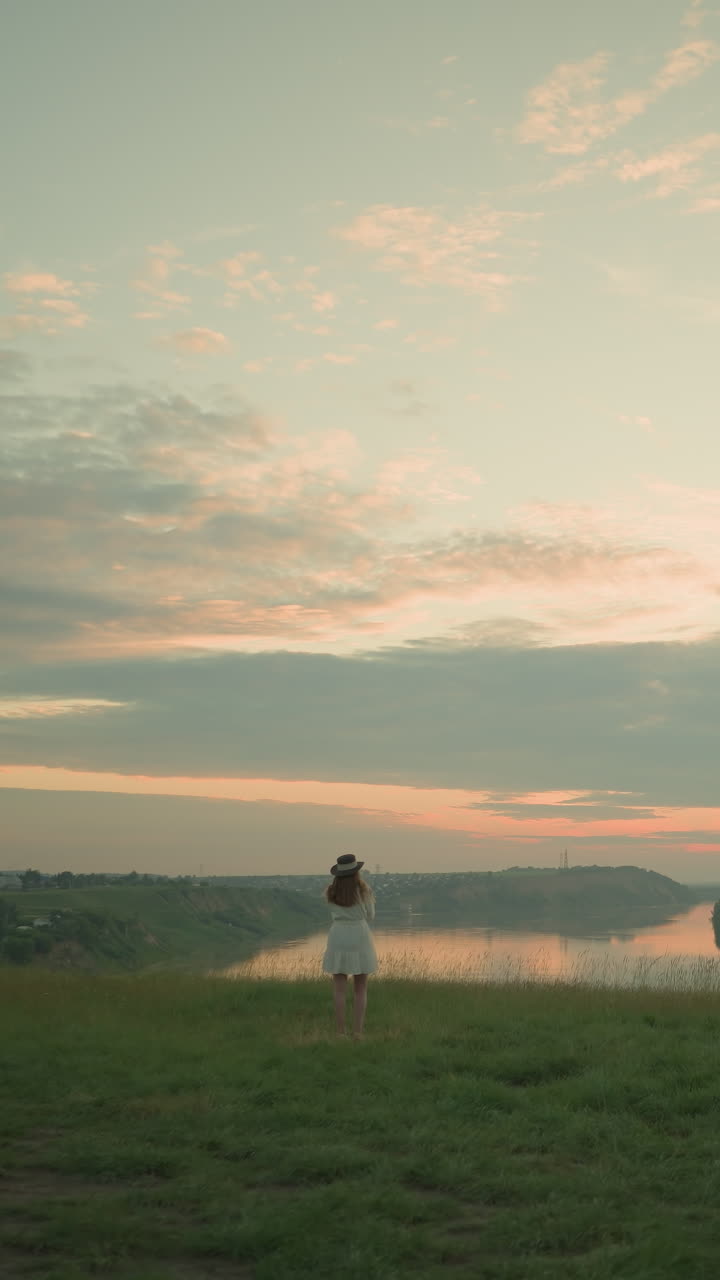 색 드레스와 모자를 입은 여자가 열린 풀에서 혼자 서서 조용한 호수와 멀리 떨어진 마을을 바라보고 있습니다.