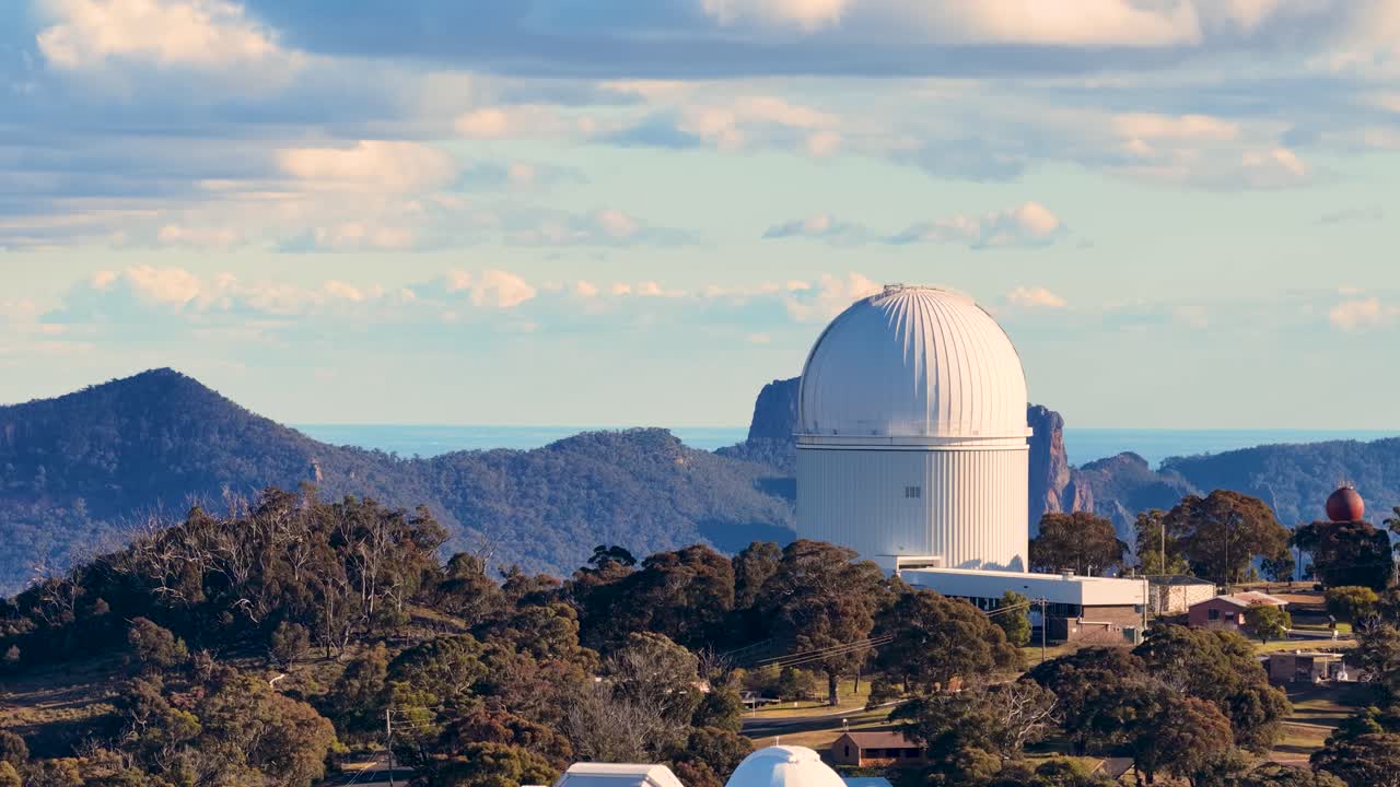 Aerial drone footage slowly pans across a large astronomical observatory dome surrounded by forested hills, under soft sunset lighting and partly cloudy skies