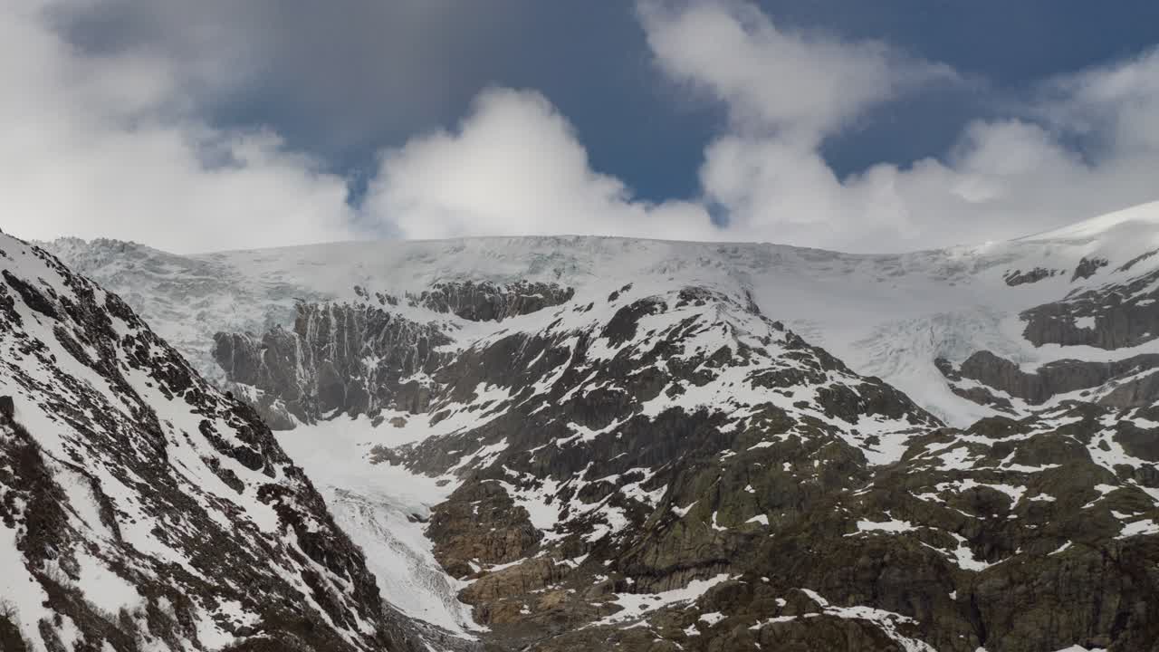 Aerial view of the Buarbreen glacier in the Folgefonna national park near Odda, Norway. Clouds crawling atop the dark snow-covered mountain, sun rays piercing through, casting flares on the glacier.