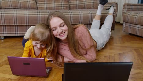 mujer niñera y niña estudiando juntos con computadora portátil, mientras yacen en el suelo cálido en casa