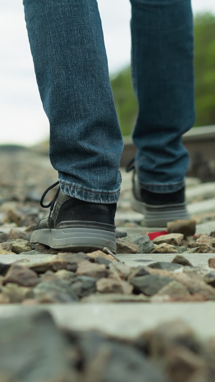 vista cercana de la pierna de un hombre en vaqueros y zapatos de lona, caminando tranquilamente por vías ferroviarias rocosas, con una vista borrosa de árboles y postes eléctricos