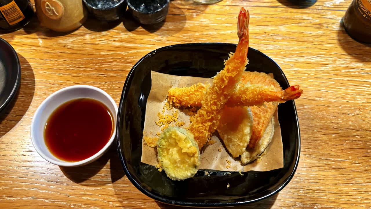Imagens em close-up de tempurá de camarão servido em uma tigela com molho de acompanhamento. Camarão frito levemente empanado e textura crocante destacam a culinária japonesa clássica e a apresentação de restaurante