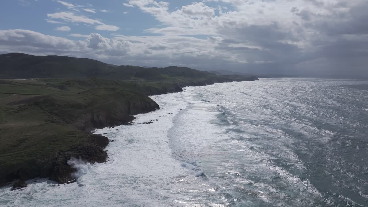 Slow orbital drone flight moving left over Santa Tecla beach in Cantabria, Spain, showing turquoise waters, rocky cliffs, green meadows, and small coastal villages under a soft cloudy summer sky