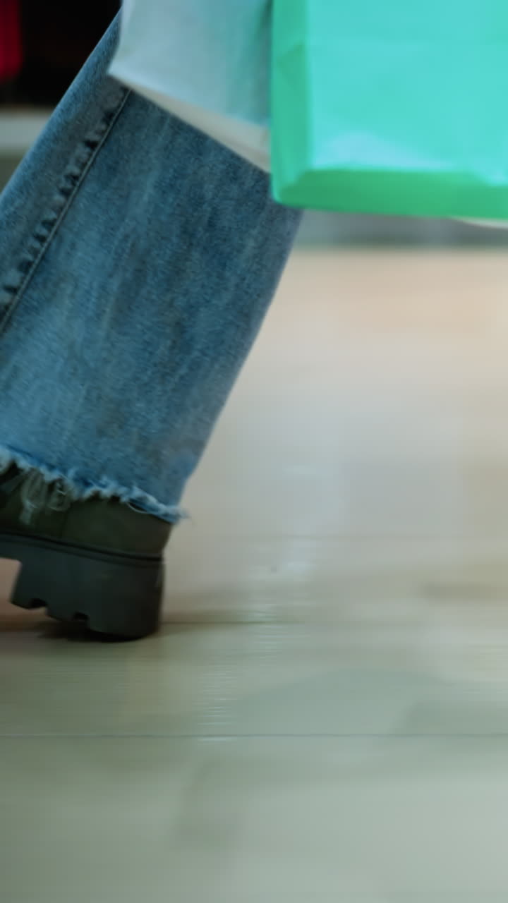 Vista laterale delle gambe di una persona in jeans sfrangiati e stivali robusti che cammina in un centro commerciale portando borse della spesa. Ombra tenue sul pavimento piastrellato e borse colorate sfocate in uno spazio commerciale luminoso e ben illuminato, viste attraverso il vetro.