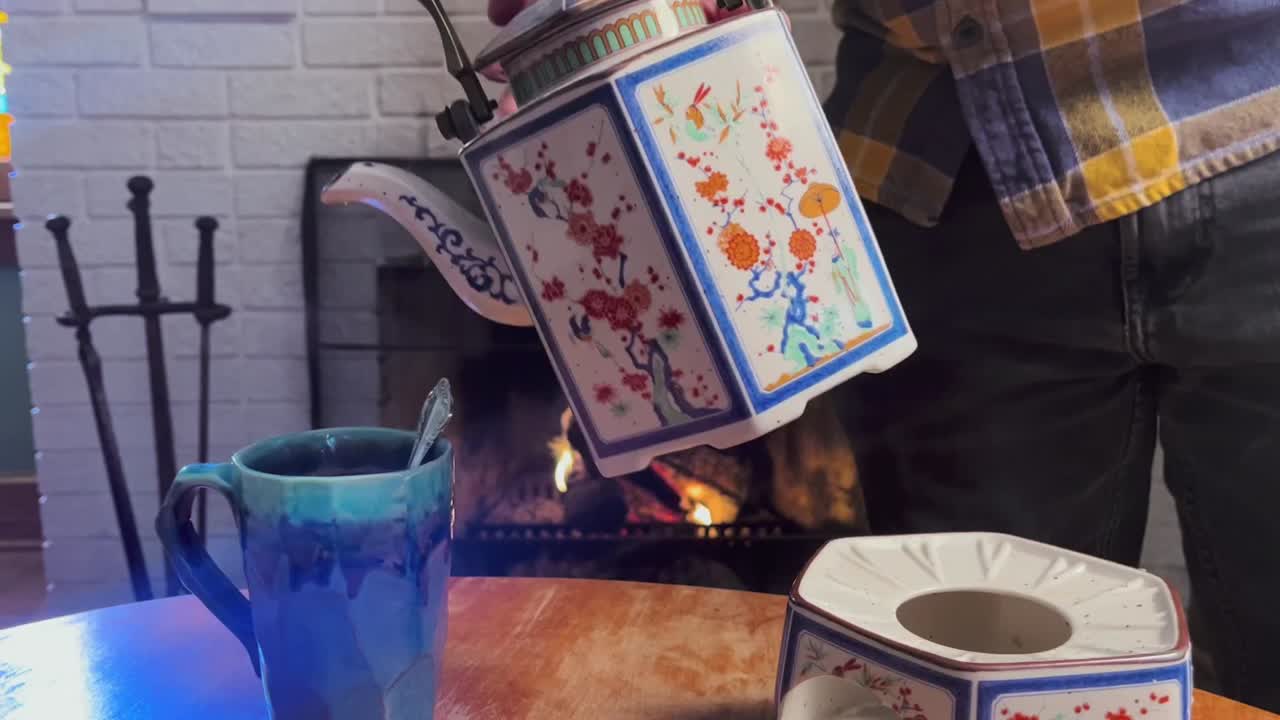 A hot cup of tea poured from an antique tea pot in front of a cozy fireplace