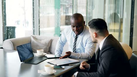 two multi-ethnic businessmen looking at diagrams and graphs on laptop screen and discussing the financial report of their startup in modern cafe during lunch time.
