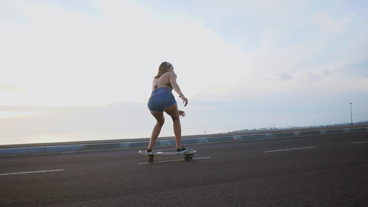 A stylish young skateboarder glides on her board in shorts along a mountain road at sunset, the view of the mountains showcased in slow-motion beauty