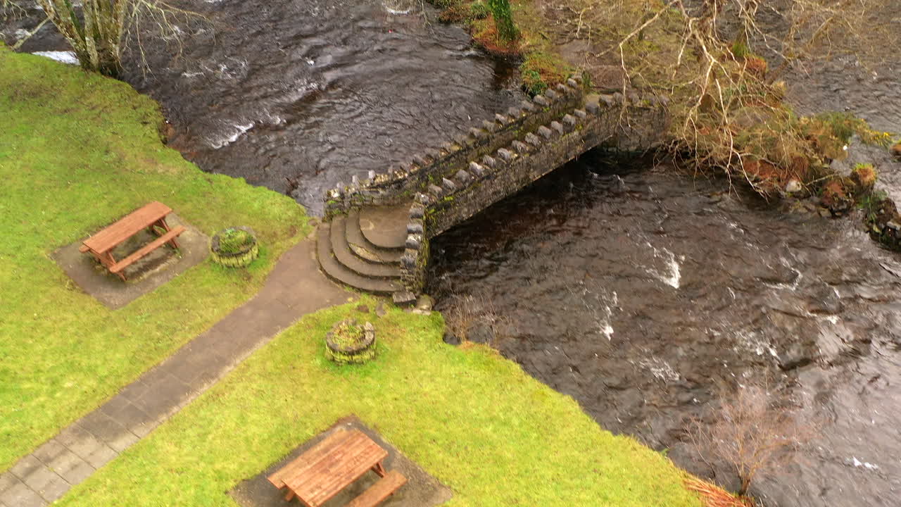 Small wooden benches sit beside a winding river and old stone bridge, surrounded by dense green trees and grass
