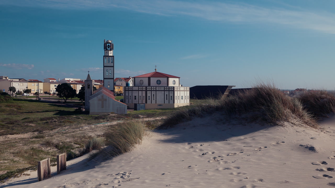 A modern white church with a distinctive striped bell tower stands among sandy coastal dunes near Aveiro, Portugal, with sea grass and blue sky creating a serene seaside atmosphere.