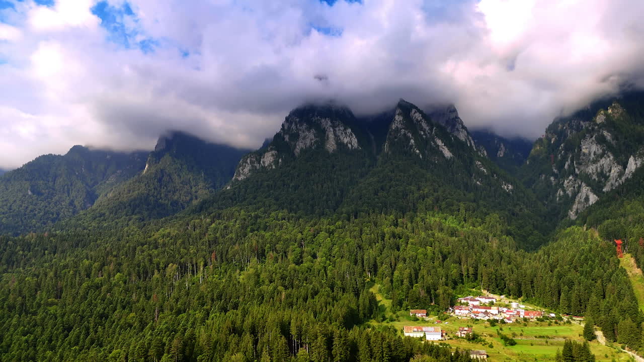 Mountains rise above forest and village in scenic landscape near winter. Clouds cover peaks of mountains above a green forest and small village during daylight