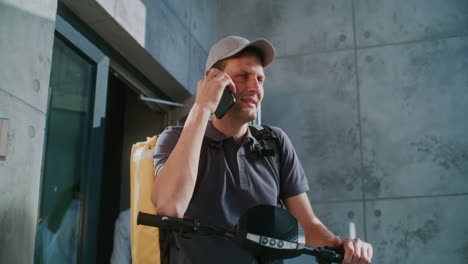 a delivery person on an e-scooter talks on a mobile phone outside a modern building