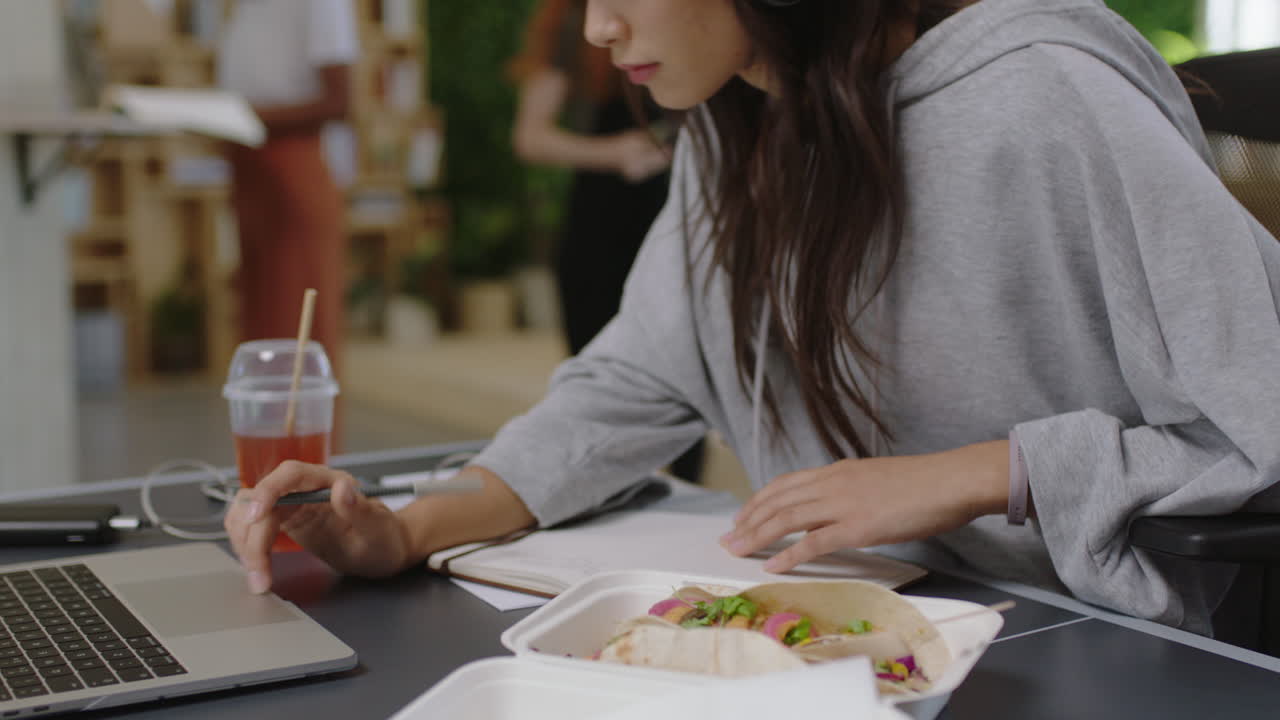 젊은 아시아 사업가 여성 메모를 쓰고 공부를 즐기고 음악을 듣고 헤드폰을 착용하는 여성 학생이 바쁜 다민족 사무실에서 일합니다.
