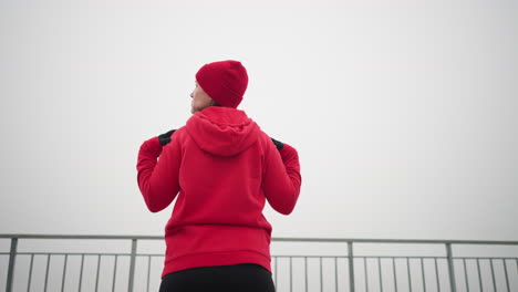 back view of woman performing hand stretch by turning her arm and head left and right, dressed in athletic wear, with iron railing and serene outdoor atmosphere in background