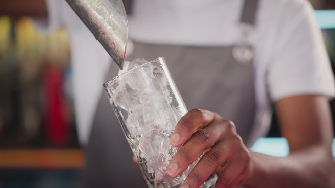 Barman pours ice pieces with scoop into glass closeup. Black bartender prepares ice cubes for refreshing cocktail. Fresh beverages for visitors in bar
