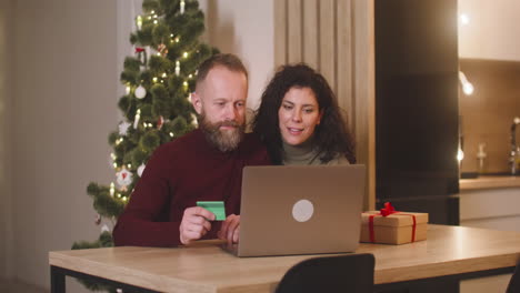 pareja comprando en línea con una tarjeta de crédito usando una computadora portátil sentada en una mesa cerca de un regalo en una habitación decorada con un árbol de navidad 1