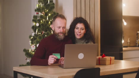 couple buying online with a credit card using a laptop sitting at a table near a present in a room decorated with a christmas tree 1