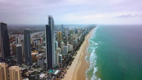 aerial forward moving view of skyscraper city by the beach with turquoise blue ocean on a cloudy day