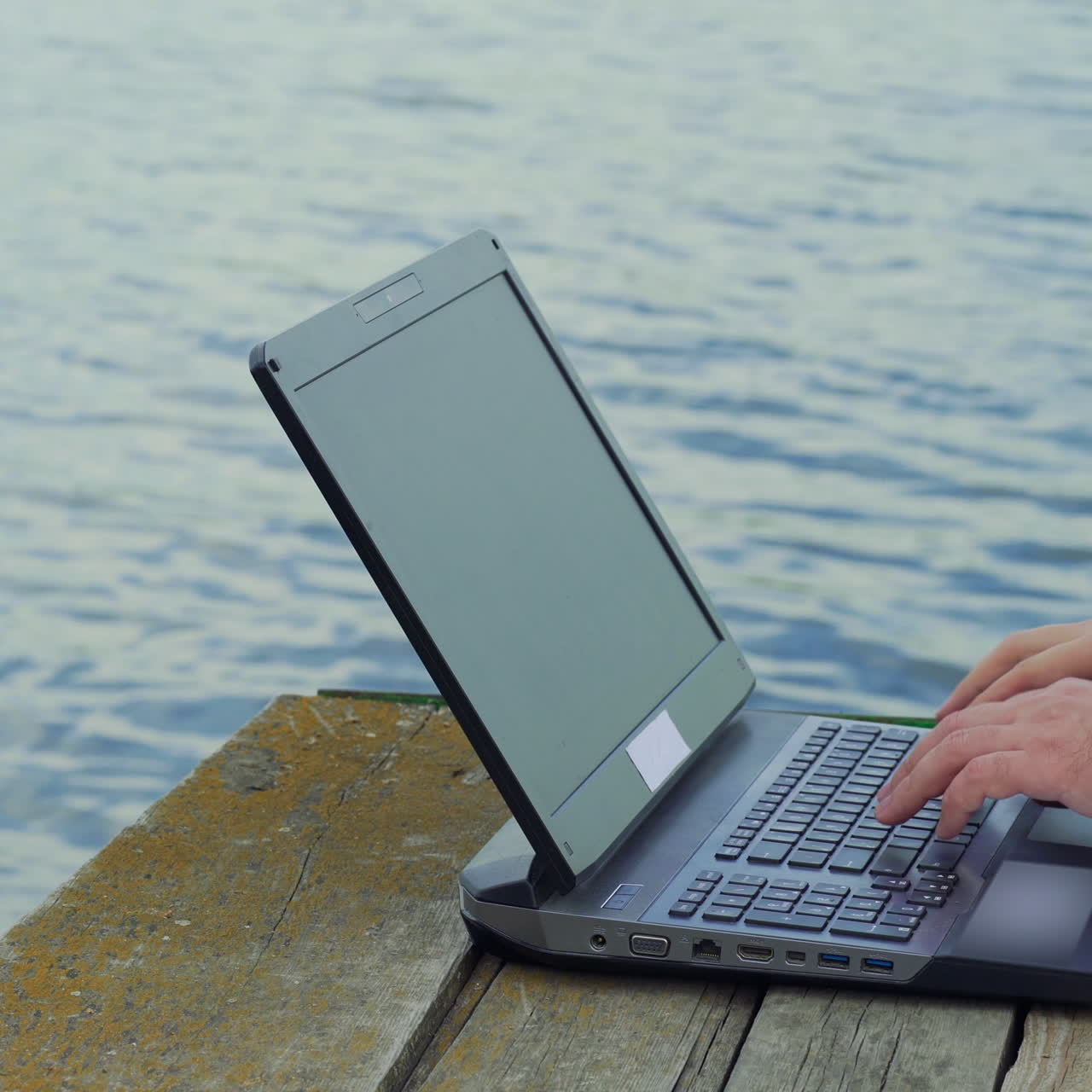 Modern laptop on the old wooden bridge and mans hands typing on it over the river background. Man working on laptop outdoors. Close-up