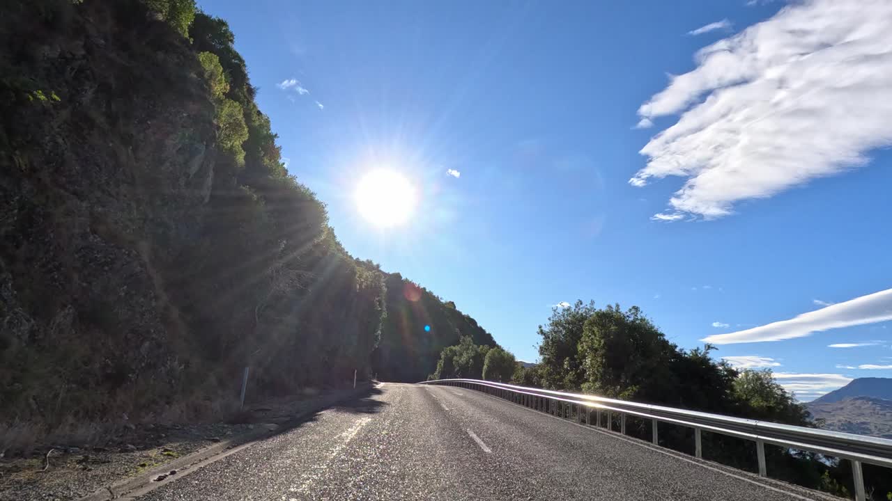 A serene drive along Queenstown's winding roads, capturing sunlit landscapes and lush greenery under a clear blue sky