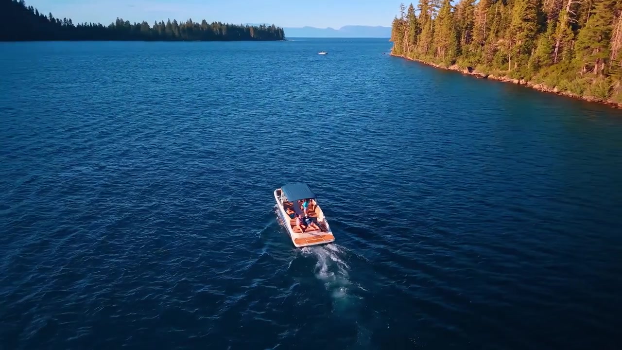 Motorboat leaving a wake across Emerald Bay. A white hull crosses blue water beside sunlit conifers, with mountains visible beyond the inlet.